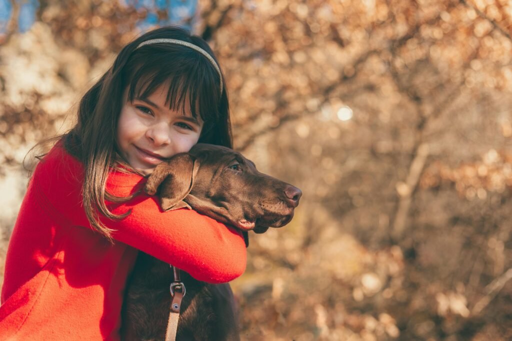 Happy girl in red hugging a chocolate Labrador in an autumn setting, showcasing warmth and affection.