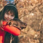 Happy girl in red hugging a chocolate Labrador in an autumn setting, showcasing warmth and affection.
